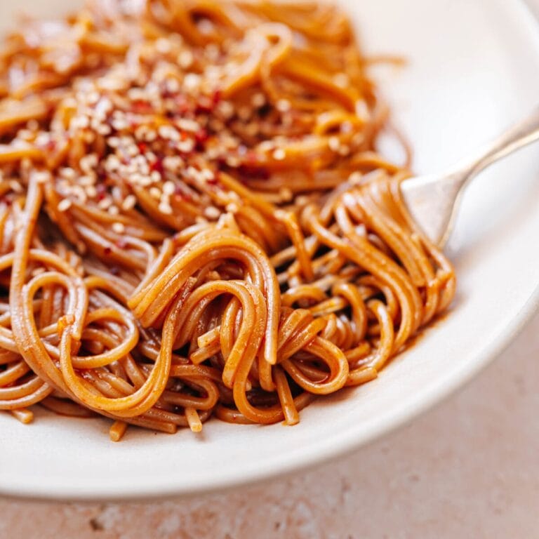 A plate of gochujang noodles topped with sesame seeds and chili flakes, with a fork resting beside them.