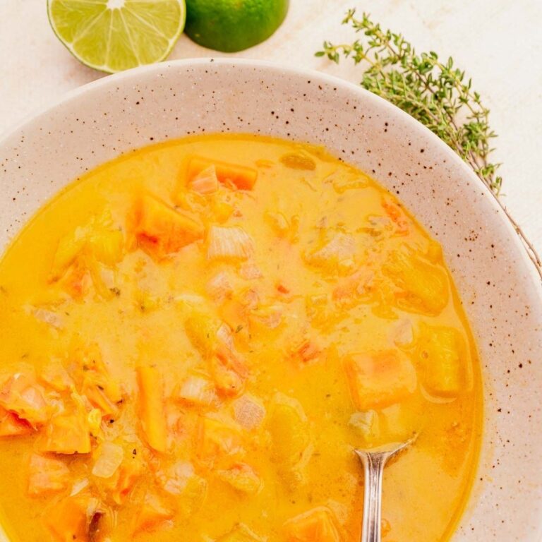 A bowl of Jamaican pumpkin soup with chunks of vegetables and a spoon. Lime halves and fresh herbs are in the background.